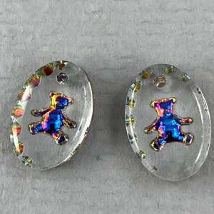 Peut inclure: Deux breloques ovales en verre transparent avec un motif d'ours en peluche bleu iridescent métallique. Les breloques ont un trou en haut pour être suspendues.