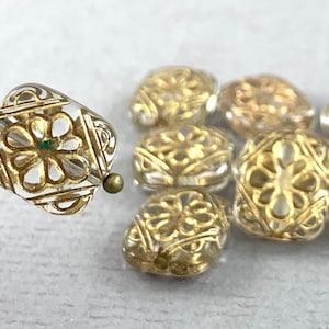May include: Close-up of several clear and gold-colored beads. One bead is on a pin, showcasing a square shape with a floral design and a small green accent. The other beads have a similar design and shape.