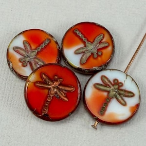 May include: Four round beads featuring a marbled orange and white glass surface with a raised metallic dragonfly motif on each. One bead is positioned on a thin metal pin, while the other three are arranged in a cluster around it.