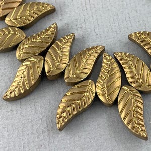 May include: A strand of gold-colored leaf-shaped beads with a textured surface. The beads are arranged in a staggered pattern.