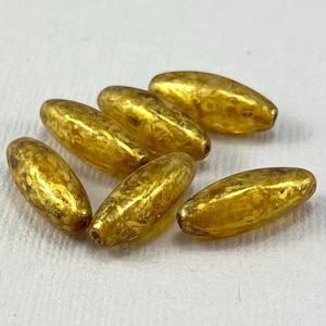 May include: Six gold-colored glass beads with a marbled pattern. The beads are oval shaped and have a glossy finish.