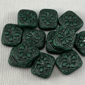 May include: A collection of dark green square beads with a carved floral design. The beads are made of glass or ceramic and have a matte finish.