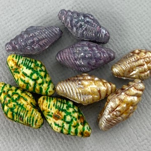 Może przedstawiać: Zestaw ośmiu szklanych koralików w różnych kolorach i wzorach. Korale mają kształt muszli i błyszczące wykończenie. Kolory obejmują fioletowy, zielony i złoty.