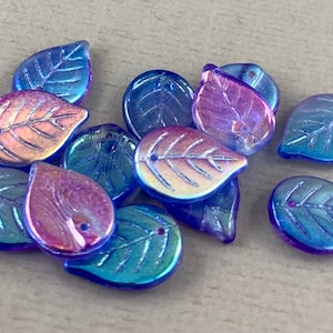 May include: A collection of iridescent leaf-shaped beads. The beads are a mix of blue, purple, and gold, with a glossy finish. Each bead has a small hole for stringing and detailed veining.