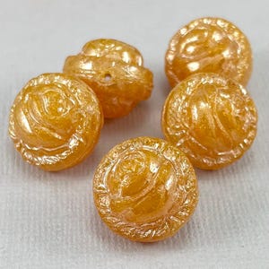 May include: Five orange-gold rose-shaped buttons are shown. The buttons have a glossy finish and a detailed floral design. The buttons are round with a raised rose in the center and a decorative border.