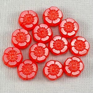 May include: A collection of red flower-shaped beads with white petal accents. The beads are arranged on a white surface, showcasing their detailed floral design. These beads are ideal for jewelry making or craft projects.