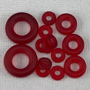 May include: A collection of red rubber O-rings of varying sizes. The O-rings are arranged on a white surface. The sizes range from small to large, with a circular shape and a hole in the center.