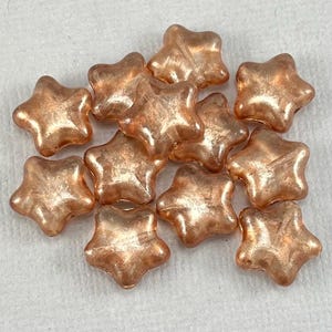 May include: A collection of copper-colored star-shaped beads. The beads have a translucent quality, allowing light to pass through. The stars are clustered together, showcasing their five-point design and reflective surface. These beads are suitable for jewelry making and crafts.