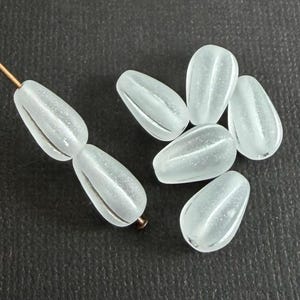 Könnte beinhalten: Eine Sammlung von mattierten Glasperlen in Tropfenform. Die durchscheinenden Perlen haben einen subtilen Glanz. Einige sind auf einem Kupferdraht aufgefädelt, andere lose.