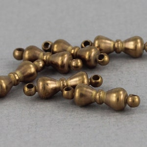 May include: Several small, antique brass-colored beads with a decorative hourglass shape. Each bead has a small loop at each end for stringing. The beads are scattered on a gray surface.
