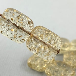 May include: Close-up of two clear, rectangular beads with gold flecks and a floral design. The beads are strung on a brown wire, with additional beads visible in the background. The beads are a decorative element.