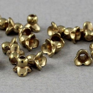 May include: A group of gold-coloured metal flower-shaped beads with a hole in the centre. The beads are scattered on a grey surface.