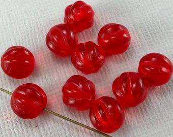 May include: A collection of ten translucent red glass beads. Each bead has a ribbed, rounded shape. The beads are scattered on a white surface, with one bead strung on a thin, gold-colored wire. These beads are suitable for jewelry making.