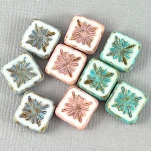 May include: A collection of square beads in various colors, including white, pink, and teal. Each bead features a raised, starburst-like design in contrasting colors. The beads have a slightly aged, vintage appearance.