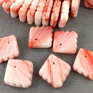 May include: A collection of pink and white shell-shaped beads with a carved design. The beads have a hole for stringing and are arranged on a gray surface. The beads are a mix of square and rectangular shapes.