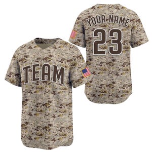 Puede incluir: Una camiseta de béisbol de camuflaje digital con la palabra "TEAM" en el pecho. La parte trasera de la camiseta muestra el número "23" y el texto "YOUR NAME". Un parche de la bandera estadounidense está en la manga.