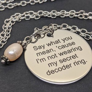 May include: A silver-toned necklace with a round pendant engraved with the text "Say what you mean, 'cause I'm not wearing my secret decoder ring." A light pink pearl and decorative beads are attached to the chain.
