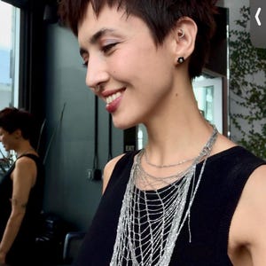 Może przedstawiać: Osoba z krótkimi, ciemnymi włosami i kolczykiem w nosie uśmiecha się, nosząc czarny top i warstwową srebrną naszyjnik. Naszyjnik ma wzór przypominający pajęczynę z zwisającymi pasmami. Tło obejmuje lustro i zieleń.