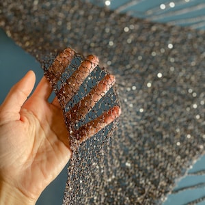 May include: Close-up of a section of black and bronze mesh fabric with sequins. The fabric is held in a hand, showcasing the intricate net-like pattern and shimmering embellishments. The background is a muted teal color.