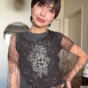 May include: Black mesh top with fringe and sparkling embellishments. The top is worn over a gray t-shirt with a skull graphic. The outfit is completed with blue jeans.