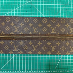 May include: Brown leather material featuring a repeating pattern of interlocking "L" and "V" letters. A brown zipper with a gold-coloured pull is centred on the material. The material is on a green cutting mat.