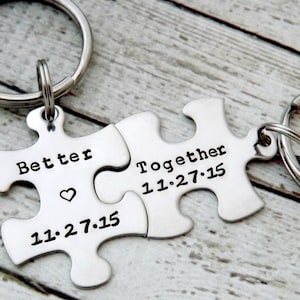 Może przedstawiać: Dwa srebrne breloki do kluczy w kształcie puzzli. Jeden element głosi "Better" z sercem i datą "11.27.15". Drugi element głosi "Together" z tą samą datą. Oba elementy są przymocowane do srebrnego breloka.