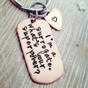 May include: A copper-colored keychain with a large rectangular tag and a small heart-shaped tag. The rectangular tag has the text "I'm a surrogate. What's your superpower?" The heart tag has a small heart symbol.