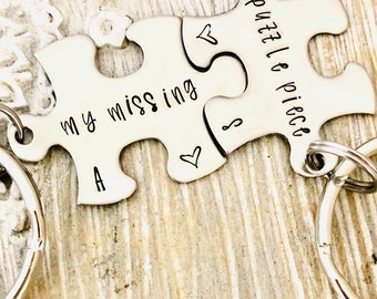 May include: Two silver puzzle piece keychains. One keychain reads "my missing A" and the other reads "puzzle piece". Each piece has a silver keyring attached. The keychains are on a light wood surface.