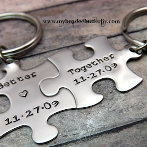 Może przedstawiać: Dwa srebrne breloki do kluczy w kształcie puzzli. Jeden element zawiera napis "Better" z sercem i datą "11.27.09". Drugi element zawiera napis "Together" i "11.27.09". Każdy brelok ma srebrny pierścień.