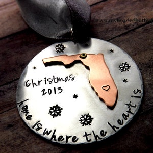 May include: A round, silver-colored Christmas ornament with a copper-colored Florida state cutout. The ornament has the text "Christmas 2013" and "home is where the heart is". Includes snowflake and star accents.