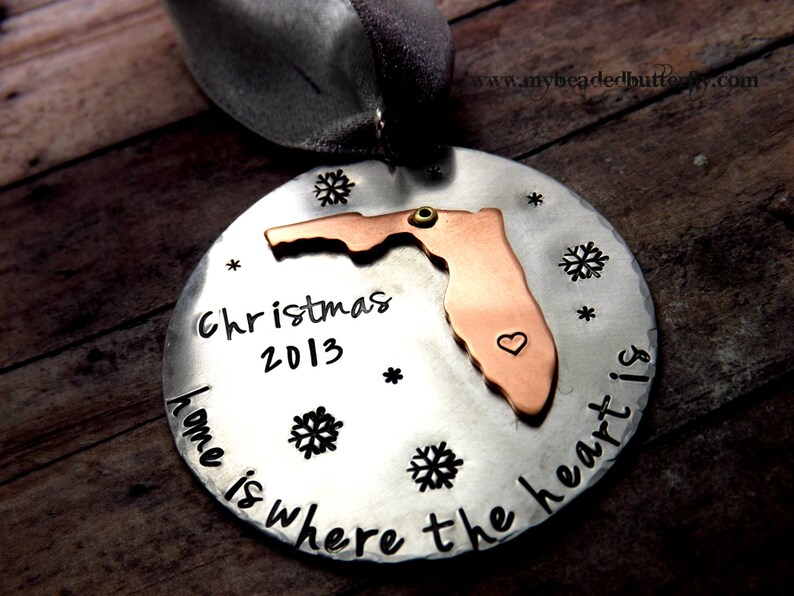 May include: A round, silver-colored Christmas ornament with a copper-colored Florida state cutout. The ornament has the text "Christmas 2013" and "home is where the heart is". Includes snowflake and star accents.