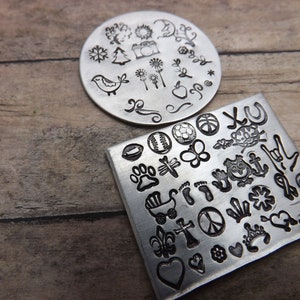 May include: Two metal stamp plates with various designs. One is round with a bird, camera, and floral elements. The other is square with a soccer ball, butterfly, owl, and other symbols. The stamps are silver.