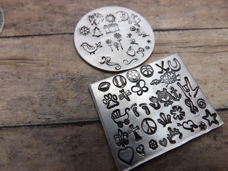 May include: Two metal stamp plates with various designs. One is round with a bird, camera, and floral elements. The other is square with a soccer ball, butterfly, owl, and other symbols. The stamps are silver.
