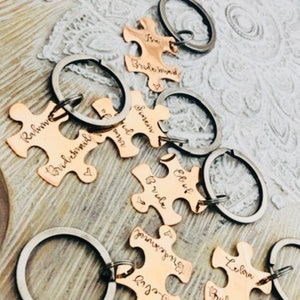 Może przedstawiać: Kolekcja breloków w kształcie puzzli w kolorze miedzi z srebrnymi kółkami. Każdy element jest grawerowany imieniem i słowem "Bridesmaid" lub "Bride". Breloki ułożone są na jasnej, teksturowanej powierzchni.