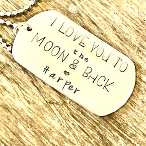 Może przedstawiać: Srebrny naszyjnik z nieśmiertelnikiem z napisem "I LOVE YOU TO THE MOON & BACK" i imieniem "Harper". Zawieszka jest na łańcuszku z koralików, na tle drewnianego tła.