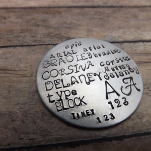 May include: A round, silver-colored metal tag with stamped text. The text includes names, words like "type" and "block", and numbers. The letters "A" and "A" are also present. The tag is on a wooden surface.