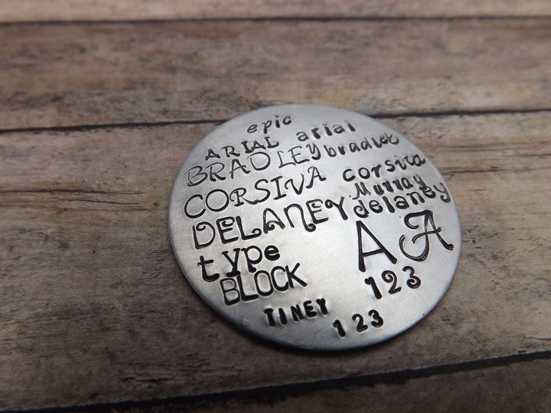 May include: A round, silver-colored metal tag with stamped text. The text includes names, words like "type" and "block", and numbers. The letters "A" and "A" are also present. The tag is on a wooden surface.