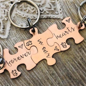 Może przedstawiać: Miedziane breloczki do kluczy w kształcie puzzli. Każdy element ma srebrny brelok i jest wygrawerowany słowami "forever in our hearts" oraz inicjałami "B". Na elementach wygrawerowane są również małe symbole serca.