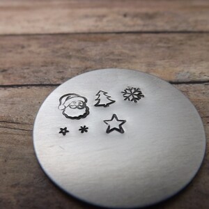May include: A silver metal disc with several Christmas-themed impressions. The impressions include Santa Claus, a Christmas tree, a snowflake, two stars, and a single star. The disc is set against a wooden background.