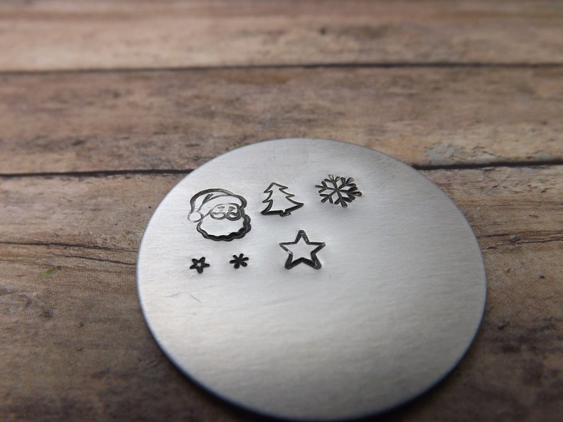 May include: A silver metal disc with several Christmas-themed impressions. The impressions include Santa Claus, a Christmas tree, a snowflake, two stars, and a single star. The disc is set against a wooden background.