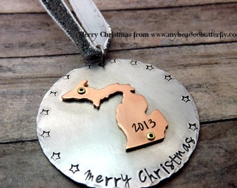 May include: A round, silver-colored Christmas ornament with a copper-colored Michigan state cutout. The ornament has a silver and white ribbon and the words "Merry Christmas" and "2013" are visible. The ornament is decorated with star shapes.