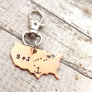 May include: Copper-colored keychain in the shape of the United States. The keychain has a silver clasp and ring. The letters "S + J" are stamped on the upper left, with dotted lines and a heart symbol. An anchor is stamped at the bottom.