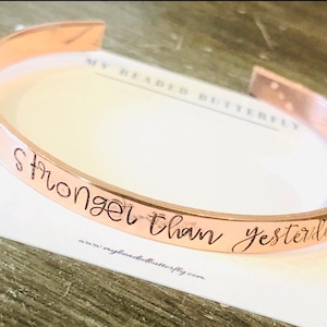 Może przedstawiać: Bransoletka typu mankiet w kolorze różowego złota z napisem "stronger than yesterday" w odręcznej czcionce. Bransoletka leży na białej karcie z tekstem "MY BEADED BUTTERFLY" i adresem strony internetowej.
