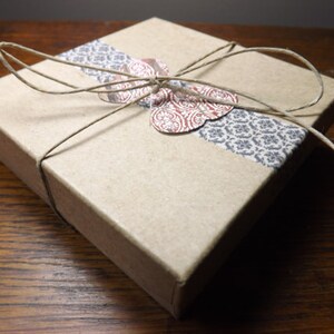 May include: A small, square, kraft paper gift box tied with twine. A decorative band of patterned paper in gray and white wraps around the box. A paper butterfly with red and white floral details is affixed to the top.