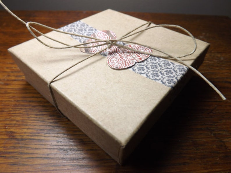 May include: A small, square, kraft paper gift box tied with twine. A decorative band of patterned paper in gray and white wraps around the box. A paper butterfly with red and white floral details is affixed to the top.