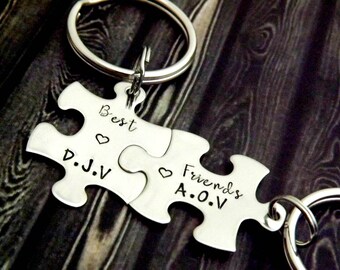 May include: A silver-colored puzzle piece keychain set. Each piece is engraved with the words "Best" and "Friends", along with initials and heart symbols. The keychain is attached to a silver ring.
