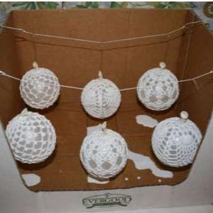 May include: A set of five spherical ornaments featuring a white crocheted lace pattern. Each ornament is suspended by a thin white string, creating a horizontal garland arrangement. The spheres have a delicate, open-work texture throughout.