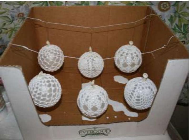 May include: A set of five spherical ornaments featuring a white crocheted lace pattern. Each ornament is suspended by a thin white string, creating a horizontal garland arrangement. The spheres have a delicate, open-work texture throughout.