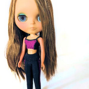 Puede incluir: Una muñeca con el pelo largo y castaño que lleva un conjunto deportivo negro y morado. La muñeca tiene ojos azules y un tono de piel claro.