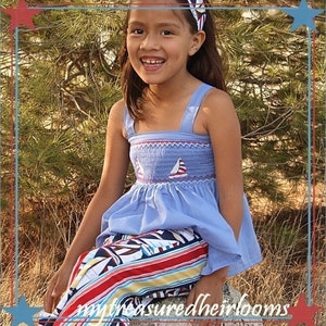 May include: A young girl wearing a blue smocked top with a white sailboat design and a red and white striped skirt. She is sitting on a rock with a green tree behind her.